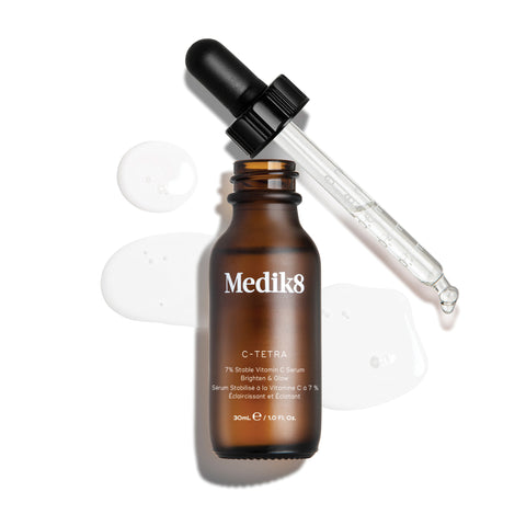 Horní „packshot“ 30ml lahvičky séra Medik8 C-Tetra Lipid Vitamin C Radiance Serum. Jantarová skleněná lahvička bez uzávěru stojí uprostřed, zatímco její černé kapátko s víčkem leží šikmo za ní. Čiré tekuté sérum je umělecky rozstříknuté a rozlité na čistém bílém pozadí kolem lahvičky, přičemž na skleněném kapátku jsou viditelné drobné kapky. Jasné osvětlení vytváří jemné stíny vlevo a dodává snímku klinický, ale zároveň elegantní vzhled.