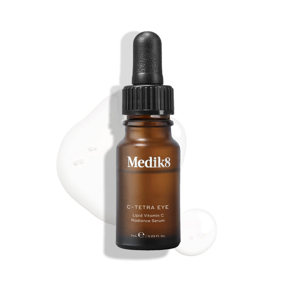 Láhev séra Medik8 C-Tetra Eye Lipid Vitamin C Radiance Serum na bílém pozadí s průsvitnými kapkami séra, které zdůrazňují jeho tekutou konzistenci.-42