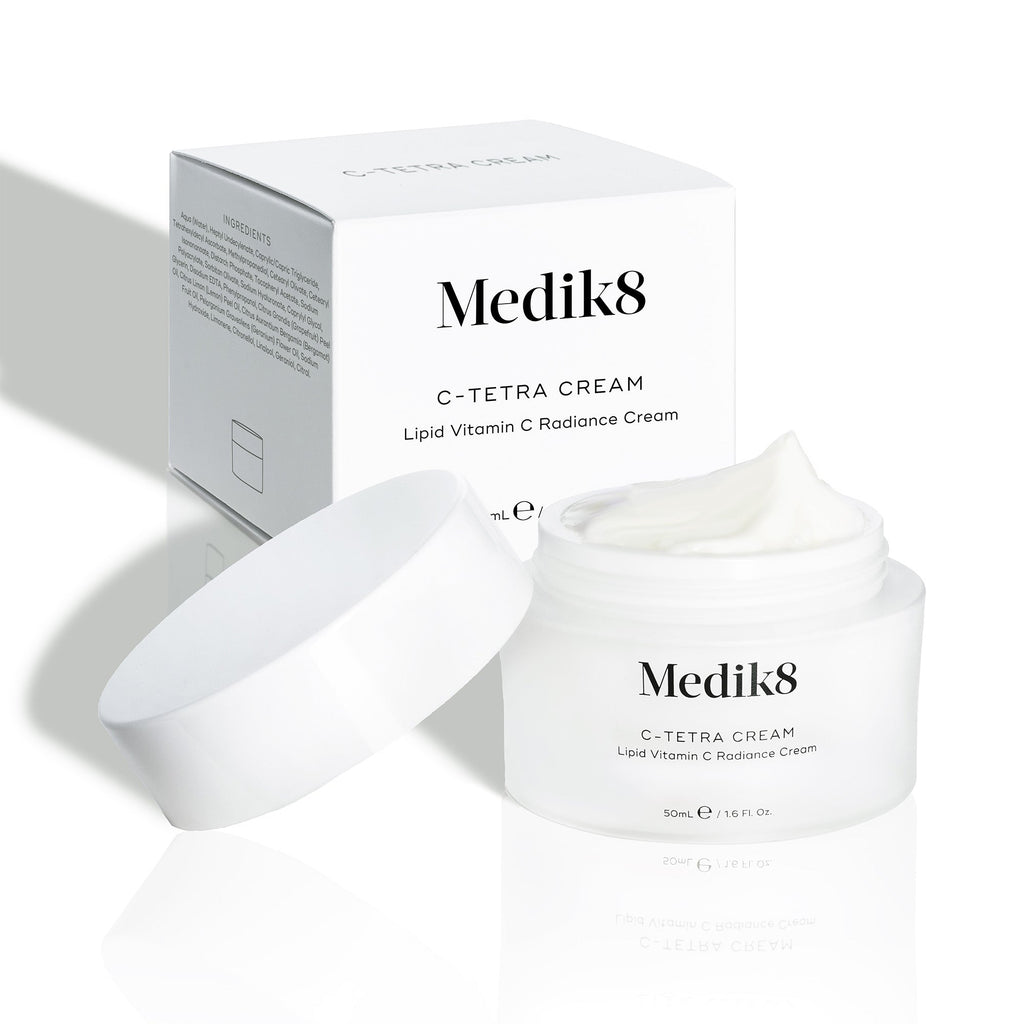Dóza Medik8 C-Tetra Cream a její bílá kartonová krabička jsou zobrazeny na čistém bílém pozadí s jemnými stíny.

Otevřená bílá matná skleněná dóza nese logo Medik8 a je označena jako C-TETRA CREAM Lipid Vitamin C Radiance Cream ve velikosti 50 ml. Na povrchu je viditelná silná vrstva bílé krémové textury. Za dózou je bílá krabička s názvem produktu a částečným seznamem složek.