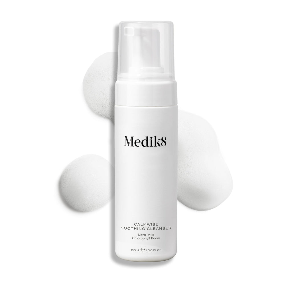 Láhev s dávkovačem čisticího přípravku Medik8 Calmwise Soothing Cleanser na bílém pozadí, za níž je vidět pruh bílé pěny, který zdůrazňuje konzistenci produktu.