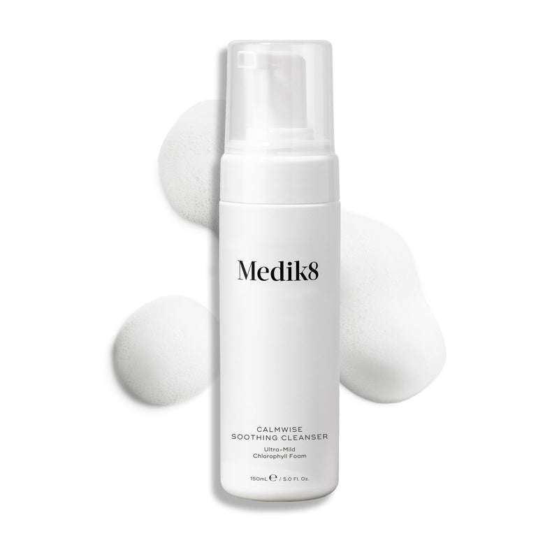 Láhev s dávkovačem čisticího přípravku Medik8 Calmwise Soothing Cleanser na bílém pozadí, za níž je vidět pruh bílé pěny, který zdůrazňuje konzistenci produktu.