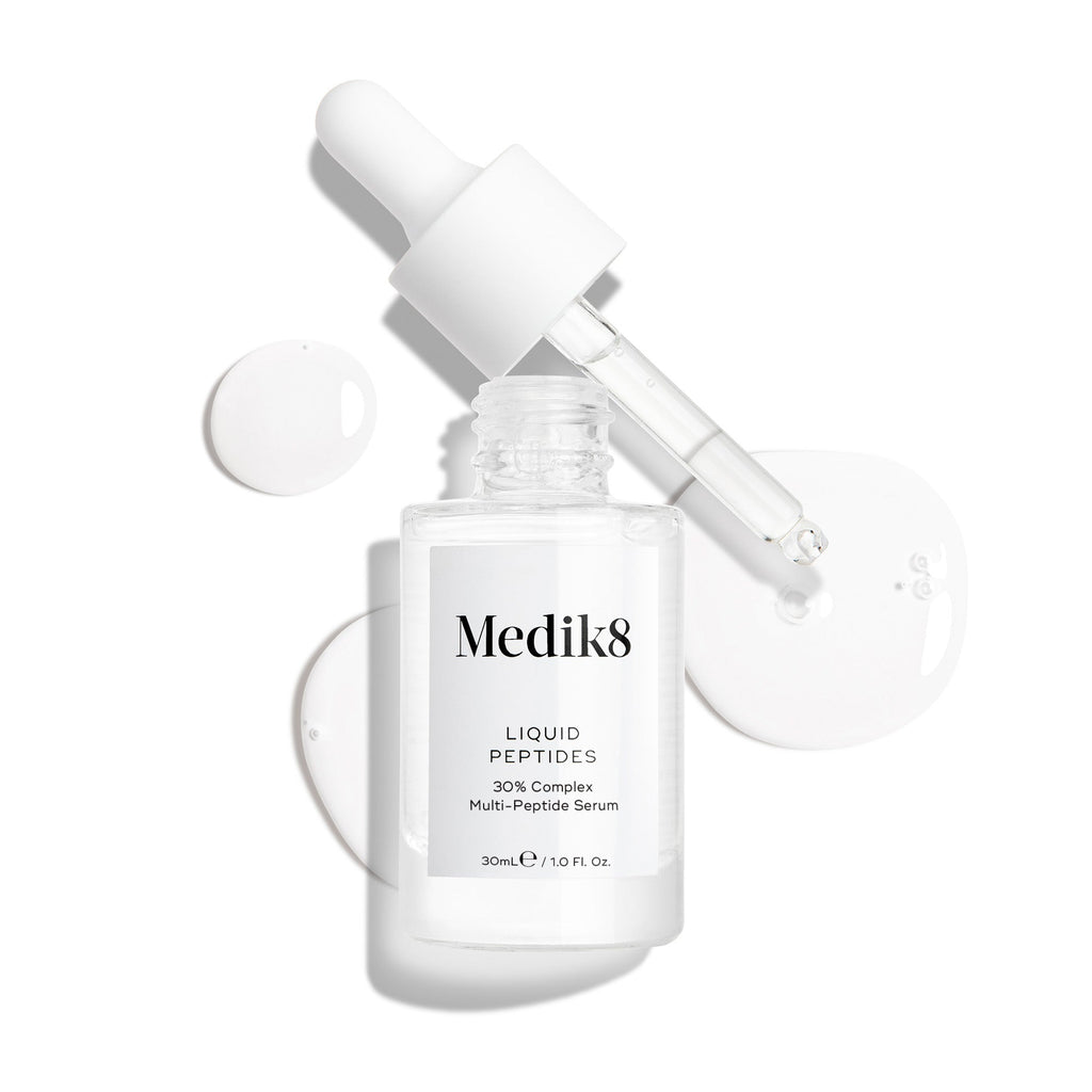 Průhledná skleněná lahvička séra Liquid Peptides je zobrazena s vyjmutým kapátkem položeným vedle ní. Kolem jsou viditelné kapky séra, které zdůrazňují jeho texturu.