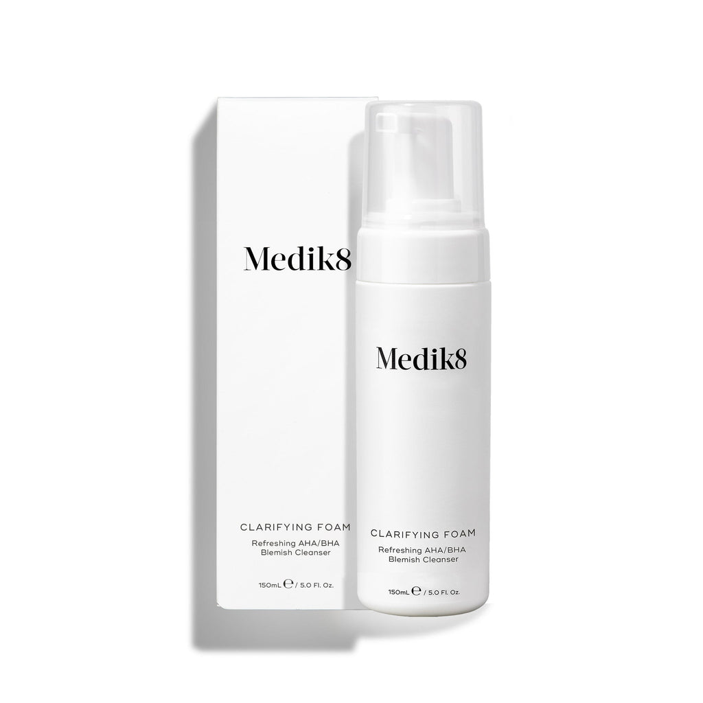 Pumpičková lahvička Medik8 Clarifying Foam stojí vedle své odpovídající bílé vnější krabičky na čistém bílém pozadí. Text na obou označuje produkt jako 150ml osvěžující AHA/BHA čisticí přípravek na nedokonalosti pleti.