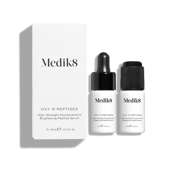 Bílá kartonová krabička produktu Medik8 Oxy-R Peptides stojí vedle dvou malých bílých lahviček séra. Jedna lahvička má černý kapátkový uzávěr, druhá má černý uzávěr.-38