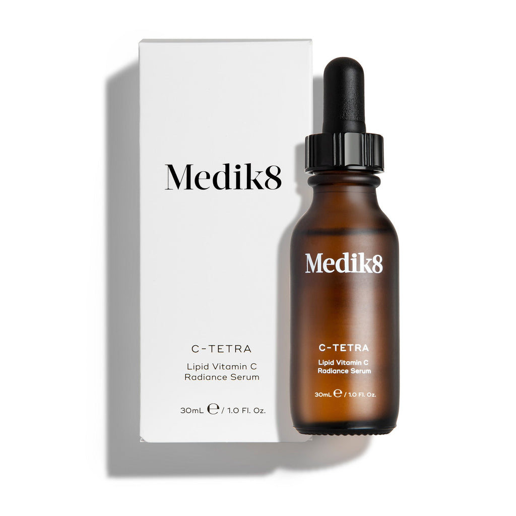 Jantarově zbarvená skleněná lahvička séra Medik8 C-Tetra Lipid Vitamin C Radiance vedle bílé kartonové krabičky. Oba produkty jsou vyfotografovány na čistém bílém pozadí s jemným stínem.