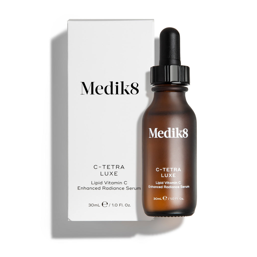 Lahvička séra Medik8 C-Tetra Luxe a její bílá kartonová krabička jsou zobrazeny na čistém bílém pozadí s jemnými stíny. Jantarově zbarvená skleněná lahvička i bílá krabička nesou logo Medik8 a označují produkt jako „C-TETRA LUXE Lipid Vitamin C Enhanced Radiance Serum“ o objemu 30 ml.