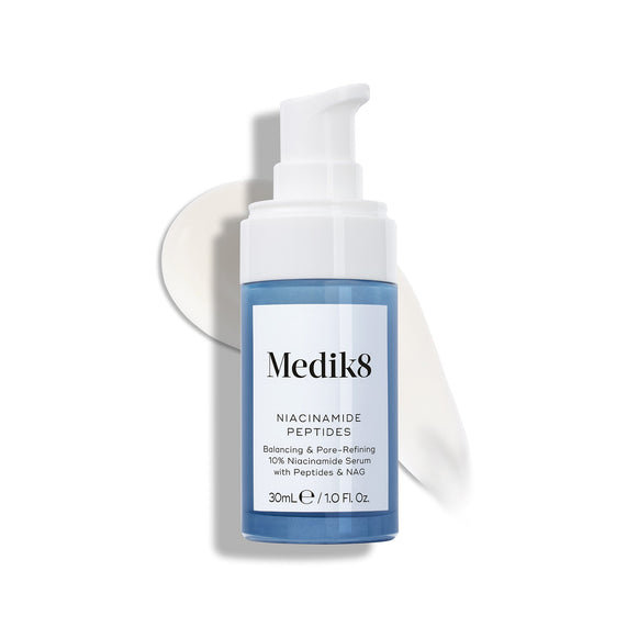 Lahvička séra Medik8 Niacinamide Peptides s bílým pumpičkovým aplikátorem je umístěna na bílém povrchu, za ní je viditelný rozetřený vzorek bílého séra.-9