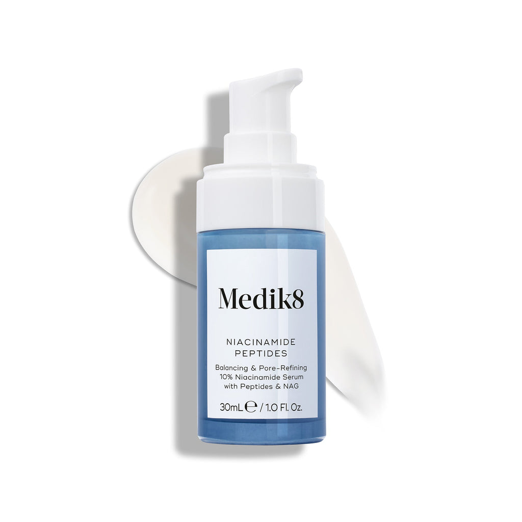 Lahvička séra Medik8 Niacinamide Peptides s bílým pumpičkovým aplikátorem je umístěna na bílém povrchu, za ní je viditelný rozetřený vzorek bílého séra.