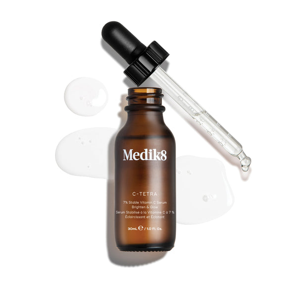 Horní „packshot“ 30ml lahvičky séra Medik8 C-Tetra Lipid Vitamin C Radiance Serum. Jantarová skleněná lahvička bez uzávěru stojí uprostřed, zatímco její černé kapátko s víčkem leží šikmo za ní. Čiré tekuté sérum je umělecky rozstříknuté a rozlité na čistém bílém pozadí kolem lahvičky, přičemž na skleněném kapátku jsou viditelné drobné kapky. Jasné osvětlení vytváří jemné stíny vlevo a dodává snímku klinický, ale zároveň elegantní vzhled.-