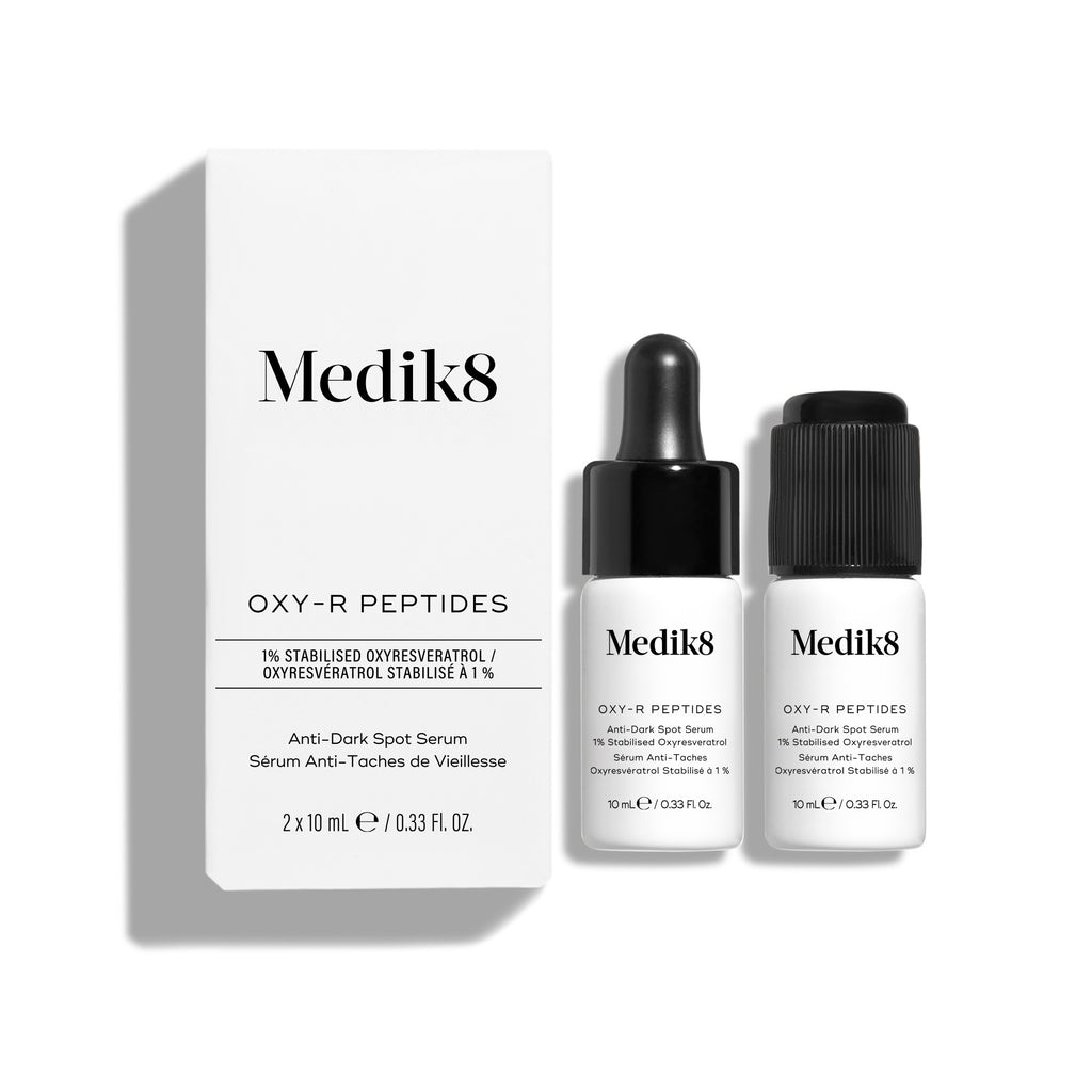 Bílá produktová krabička Medik8 Oxy-R Peptides stojí vedle dvou malých bílých lahviček séra. Jedna lahvička má černé kapátko, druhá černý uzávěr.